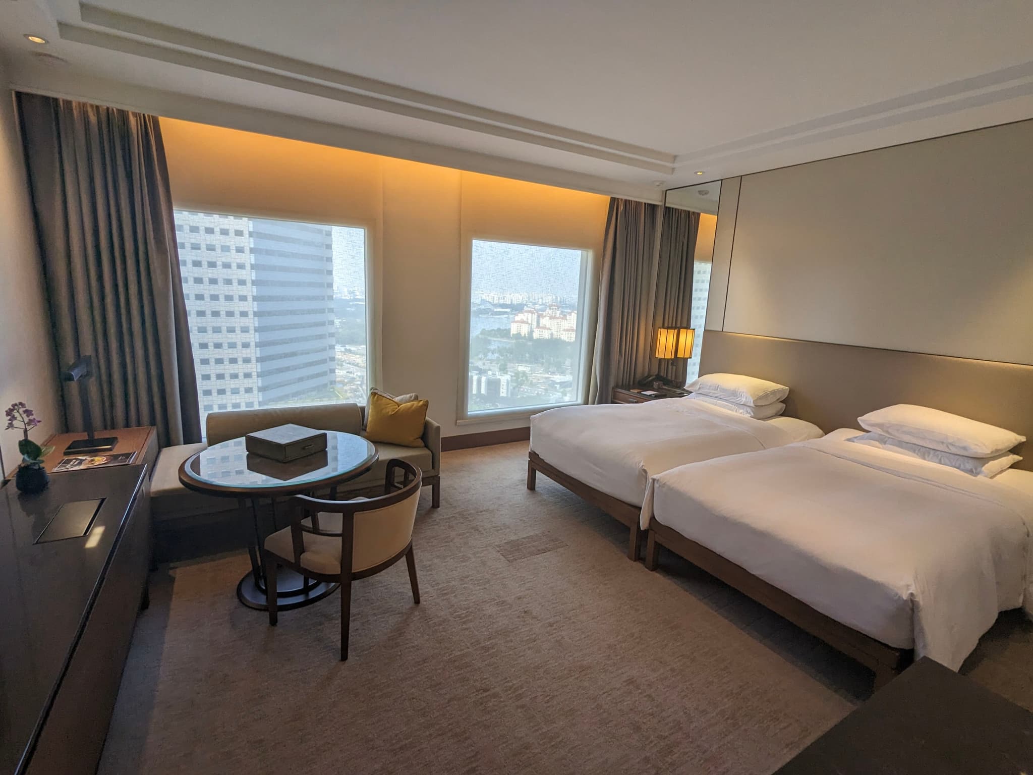 A modern hotel room with two single beds, a small round table with chairs, a desk, and large windows offering a city view