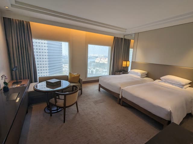 A modern hotel room with two single beds, a small round table with chairs, a desk, and large windows offering a city view