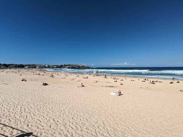 A sandy beach with scattered people sunbathing and enjoying the ocean under a clear blue sky
