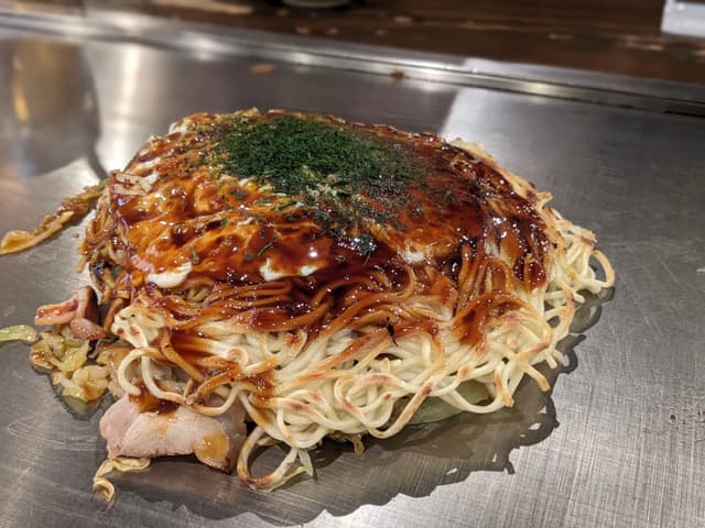 A savory Japanese pancake, likely okonomiyaki, topped with sauce and green seasoning, featuring layers of noodles and other ingredients, cooked on a griddle