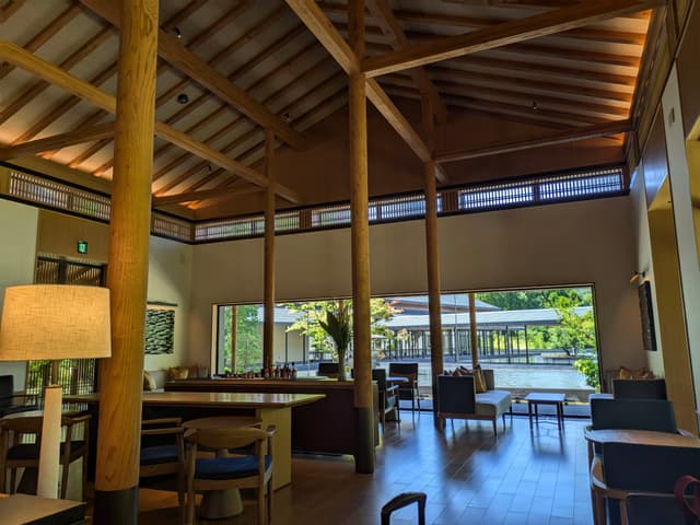 A spacious, modern interior with wooden beams, high ceilings, and large windows offering a view of greenery outside. The room features comfortable seating and a warm, inviting atmosphere