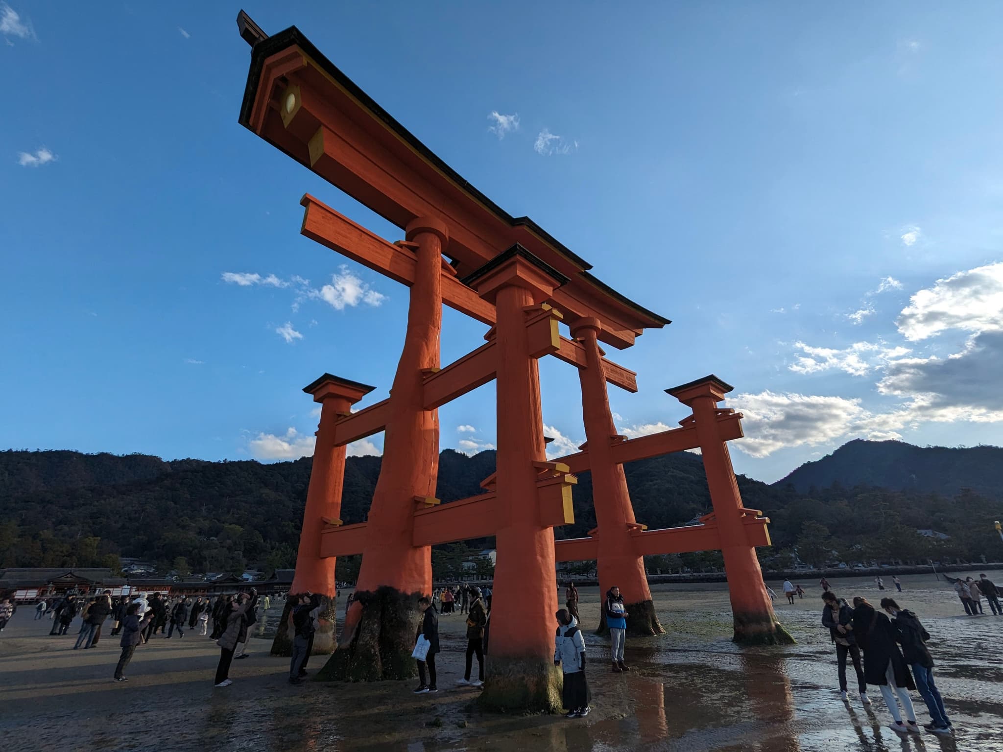A large, traditional Japanese torii gate stands in shallow water with people walking around it. The sky is clear with a few clouds, and mountains are visible in the background