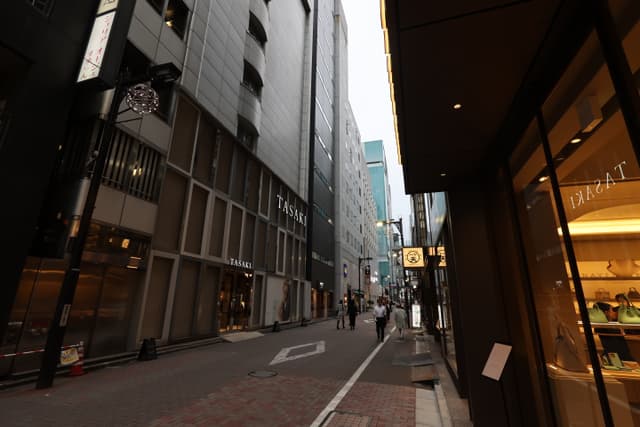 A narrow urban street lined with tall buildings, featuring a few pedestrians and a shop with a lit interior on the right