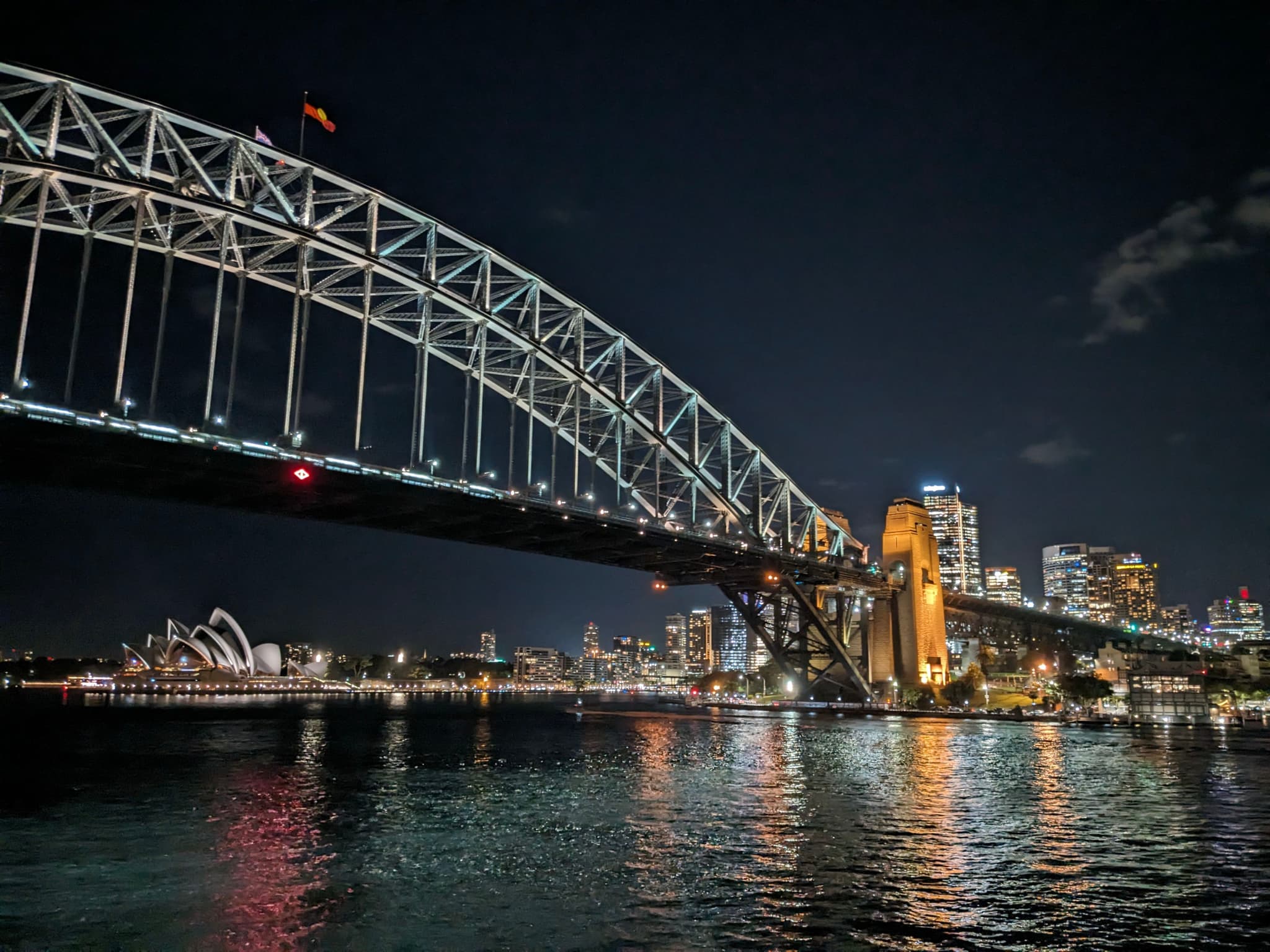 A nighttime view of the Sydney Harbour Bridge with city lights reflecting on the water and the Sydney Opera House visible in the background