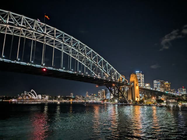 A nighttime view of the Sydney Harbour Bridge with city lights reflecting on the water and the Sydney Opera House visible in the background