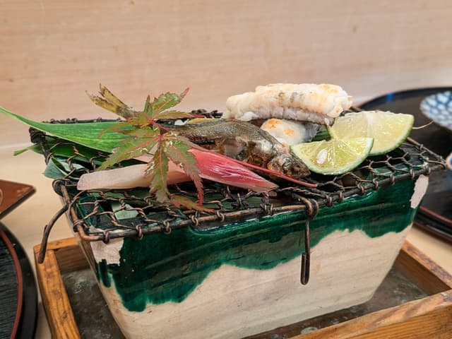 A decorative ceramic grill with assorted food items, including lime wedges, a leaf, and garnishes, arranged on top