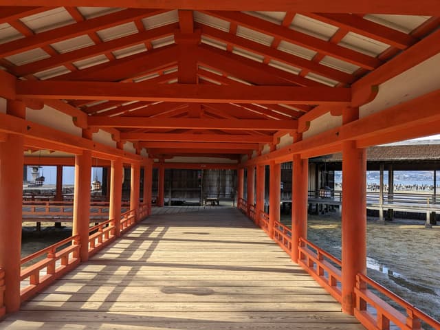 A wooden corridor with a red-painted structure and a traditional Japanese architectural style, overlooking water and distant buildings
