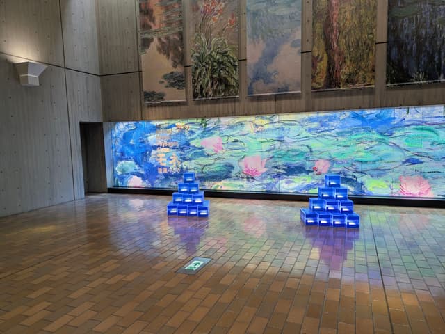 A room with a large, colorful mural on the wall, featuring abstract patterns. Two pyramid-shaped stacks of blue boxes are placed on the floor, reflecting light. The floor is made of brown tiles, and there are decorative panels above the mural