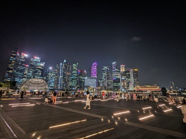 A nighttime cityscape with illuminated skyscrapers and a bustling waterfront area, featuring people walking and relaxing on a boardwalk