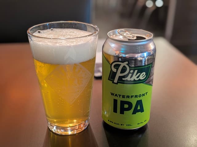 A glass of beer with a frothy head next to a can labeled Pike Waterfront IPA on a table