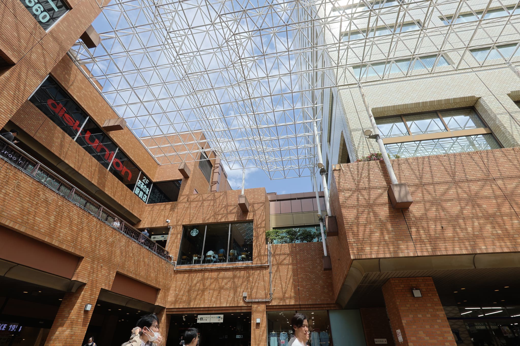 A modern architectural space with a glass ceiling, featuring multiple levels and brown brick walls People are visible in the lower part of the image, and there are signs with text on the upper levels