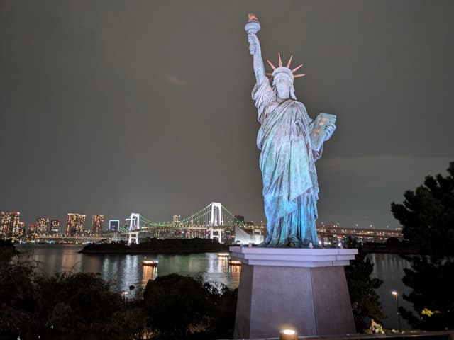 A replica of the Statue of Liberty is illuminated at night, with a cityscape and a bridge in the background