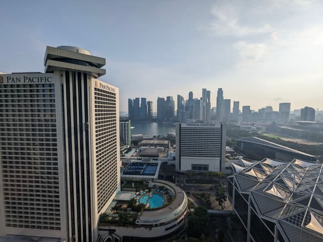 A cityscape featuring tall buildings, a large hotel with a rooftop pool, and a view of a waterfront with a skyline in the background