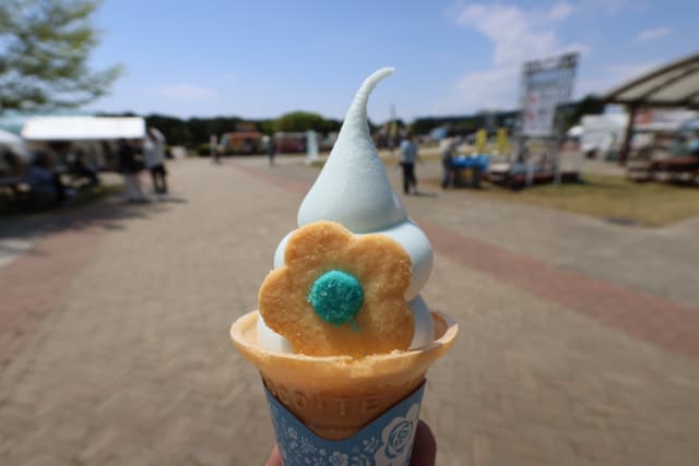 A soft-serve ice cream cone with a blue swirl and a flower-shaped cookie, held outdoors in a park-like setting with people and tents in the background