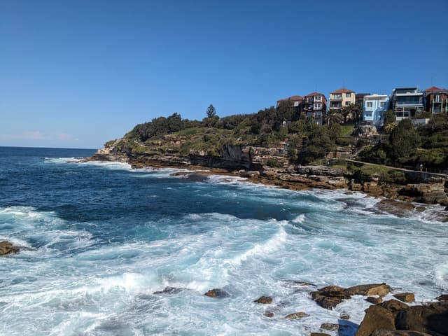 A coastal scene with rocky cliffs, waves crashing against the shore, and houses perched on the hillside under a clear blue sky