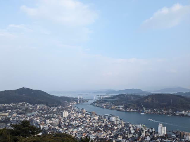 A scenic view of a coastal city with a river running through it, surrounded by hills and under a partly cloudy sky