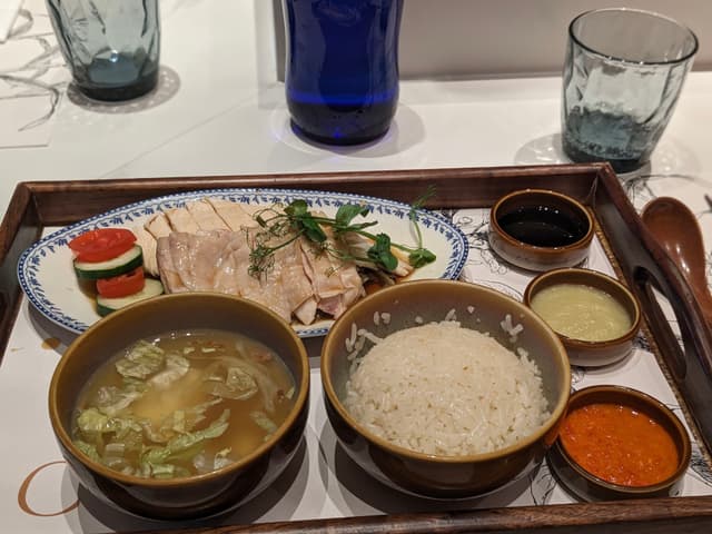 A meal tray featuring sliced chicken with garnish, a bowl of rice, a bowl of soup, and three small bowls of sauces or condiments