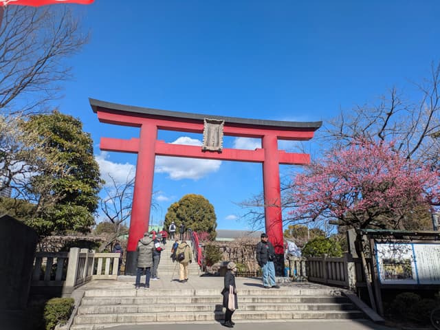 A large red torii gate stands prominently against a clear blue sky, surrounded by trees and people walking nearby
