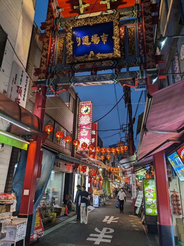 A vibrant street scene in a Chinatown area, featuring colorful lanterns, illuminated signs, and people walking along a narrow alley lined with shops