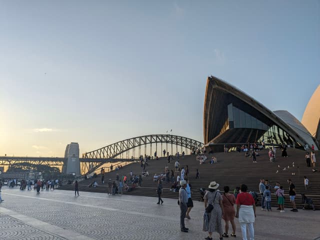 The Sydney Opera House with the Sydney Harbour Bridge in the background, surrounded by people walking and gathering on a sunny day