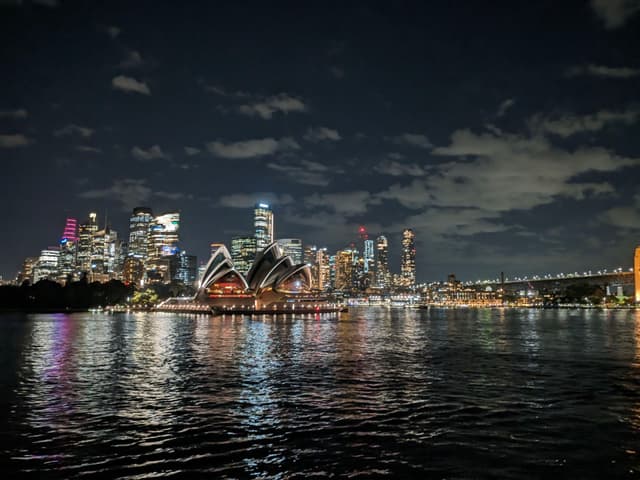 A nighttime cityscape featuring illuminated skyscrapers, the Sydney Opera House, and the Sydney Harbour Bridge, with reflections on the water