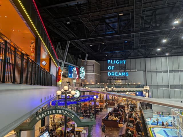 An indoor shopping and dining area with a high ceiling, featuring signs for Flight of Dreams and Seattle Terrace, and various shops and restaurants