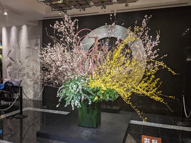 A large, artistic floral arrangement featuring pink and yellow blossoms with green foliage, set against a dark background with circular metallic elements