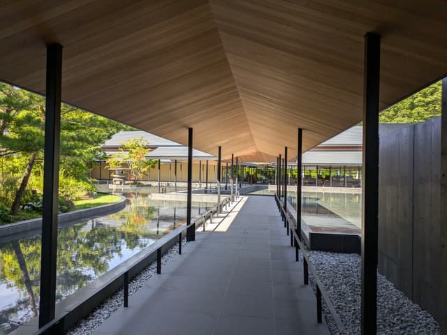A modern architectural walkway with a wooden ceiling, flanked by shallow pools and surrounded by greenery