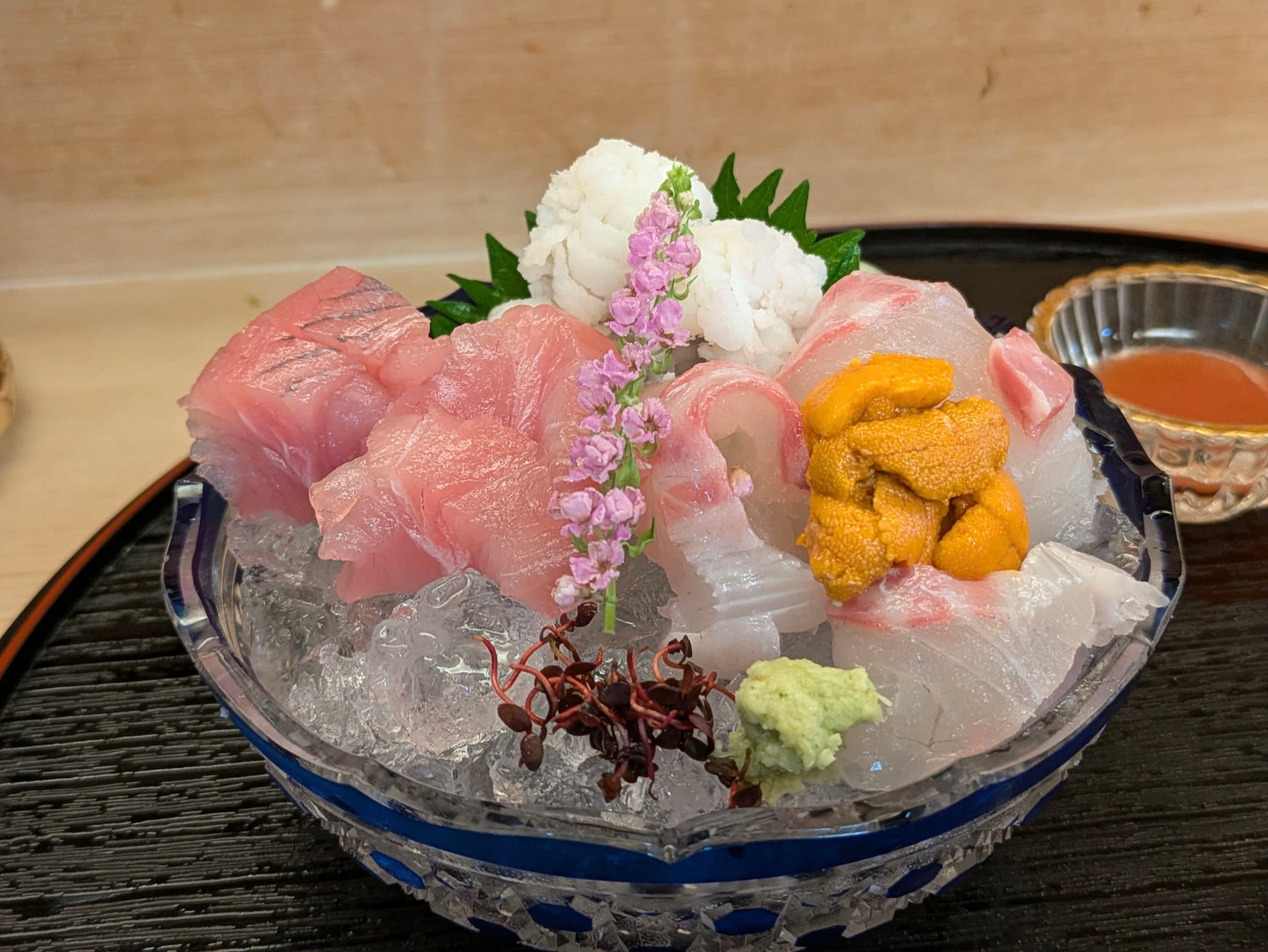 A bowl of assorted sashimi on ice, featuring various types of raw fish, sea urchin, and garnishes, with a small dish of sauce in the background