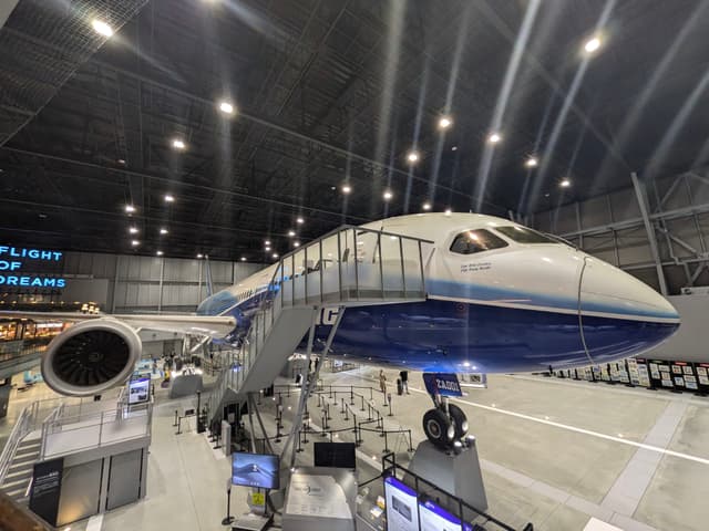 A large aircraft displayed indoors with a staircase leading to its entrance, surrounded by informational exhibits and lighting from above