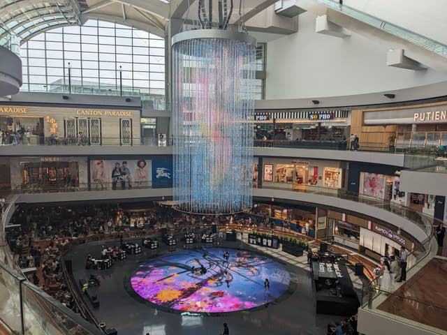 A large, multi-level shopping mall with a central circular area featuring a colorful digital display on the floor and a suspended light installation above. Shoppers are visible on different levels, and the space is well-lit with natural light from the glass ceiling