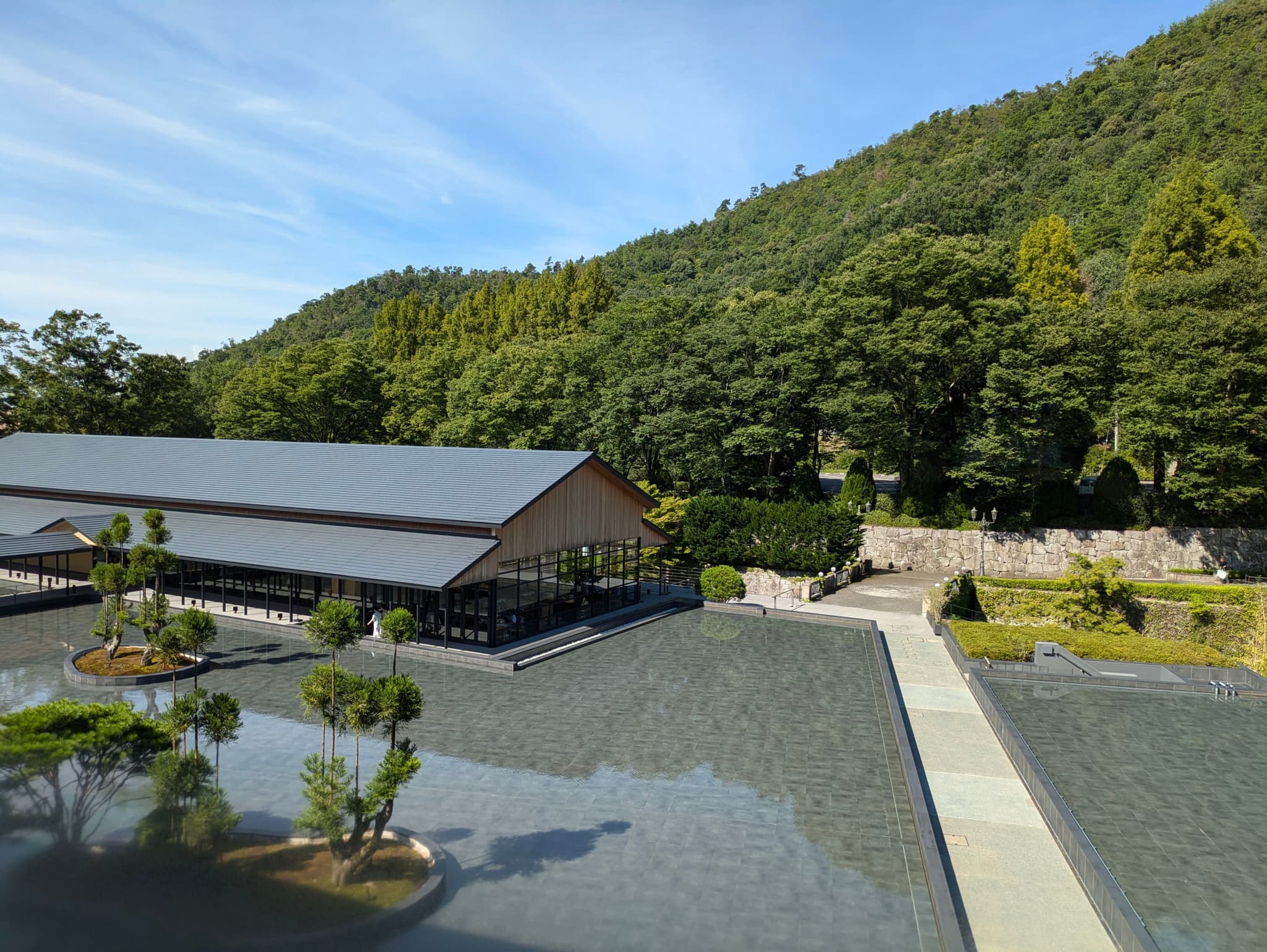 A serene landscape featuring a modern building with a sloped roof, surrounded by a reflective water surface and small trees, set against a backdrop of lush green hills and a clear blue sky