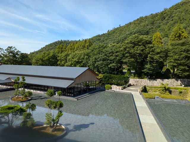 A serene landscape featuring a modern building with a sloped roof, surrounded by a reflective water surface and small trees, set against a backdrop of lush green hills and a clear blue sky