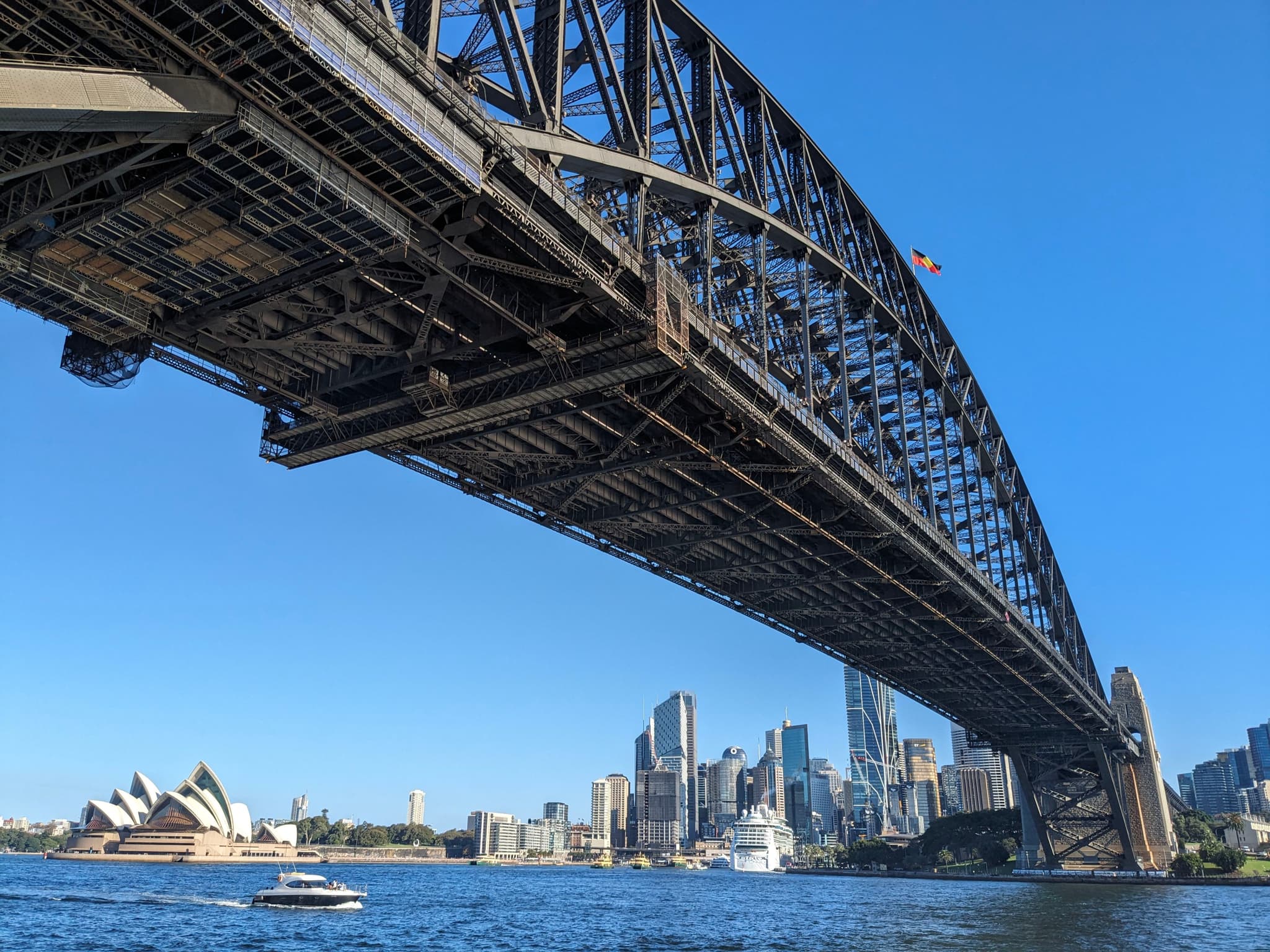 A large steel arch bridge spans across a body of water with a city skyline and a distinctive opera house in the background under a clear blue sky