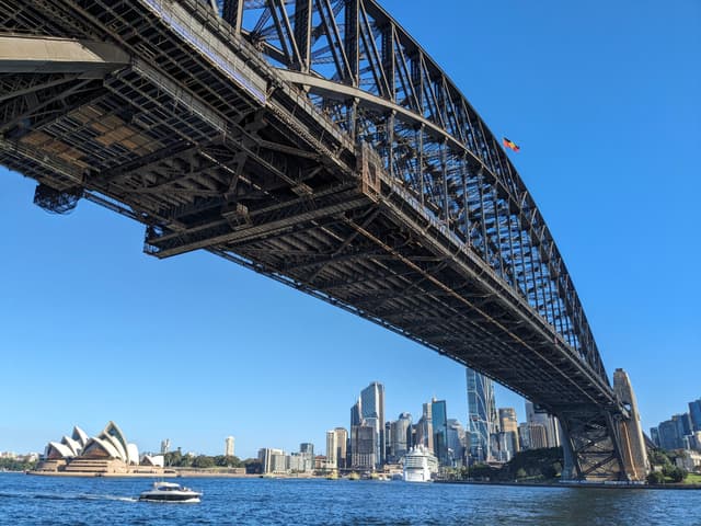 A large steel arch bridge spans across a body of water with a city skyline and a distinctive opera house in the background under a clear blue sky