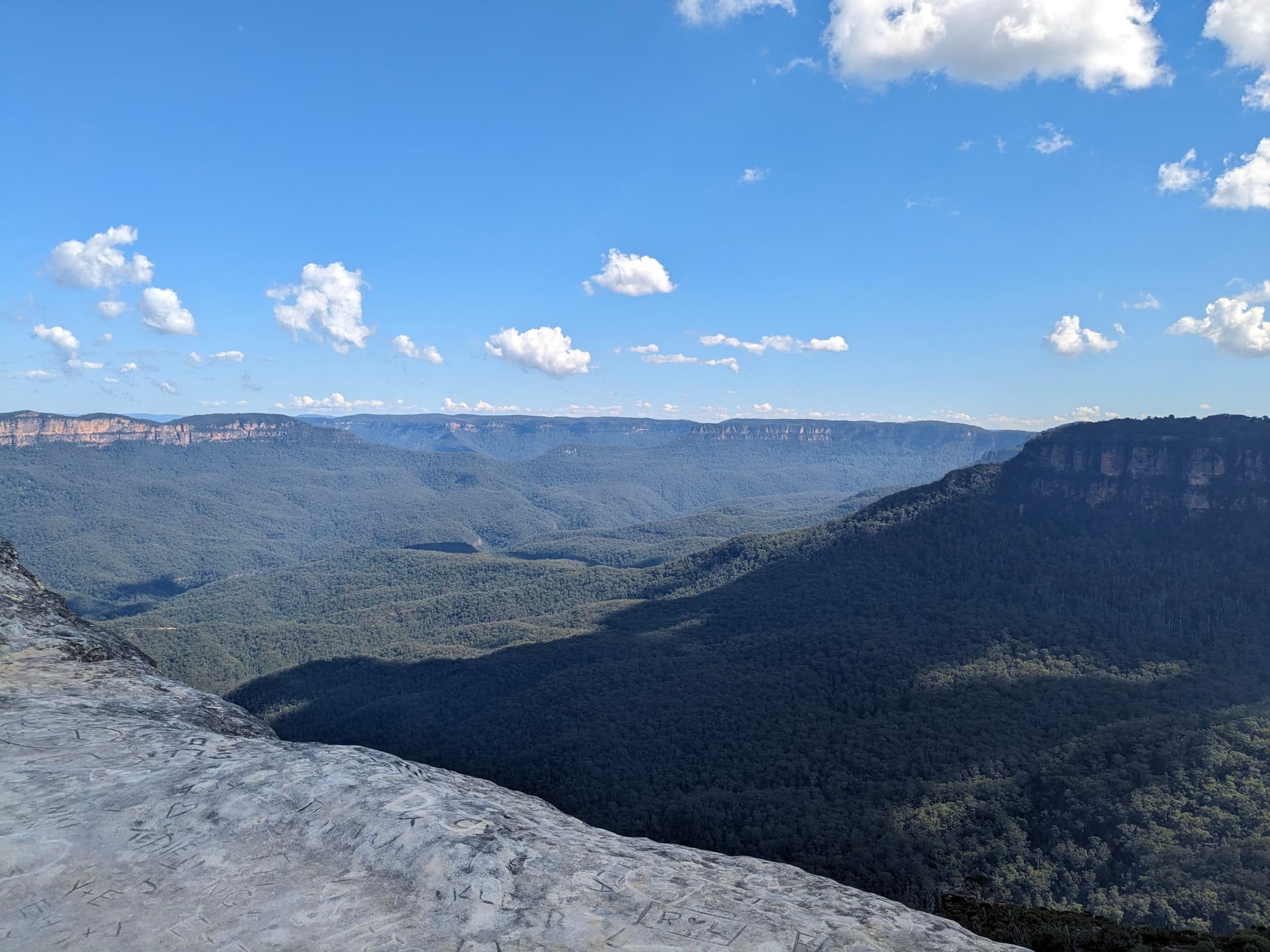 A vast landscape with a rocky foreground, expansive green valleys, and distant mountain ranges under a clear blue sky with scattered clouds