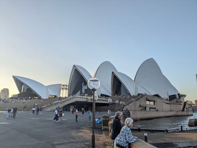 The Sydney Opera House with its iconic sail-like design, people walking nearby, and a clear sky in the background