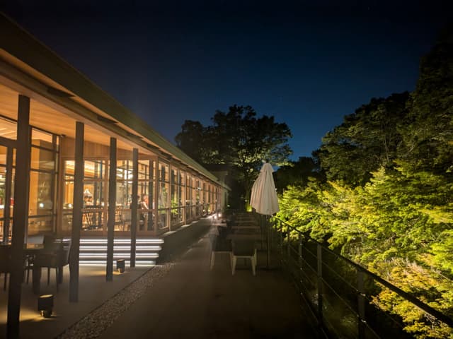 A modern building with large glass windows is illuminated at night, adjacent to a patio area with seating and surrounded by trees