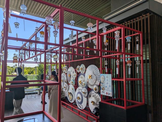 A red metal framework displaying various decorative plates and hanging ornaments, with people observing the exhibit in an outdoor setting