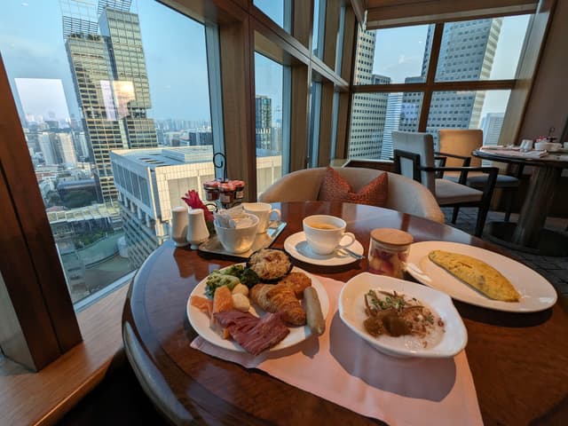 A table set with a variety of breakfast foods, including an omelet, pastries, and coffee, in a high-rise restaurant with large windows offering a cityscape view