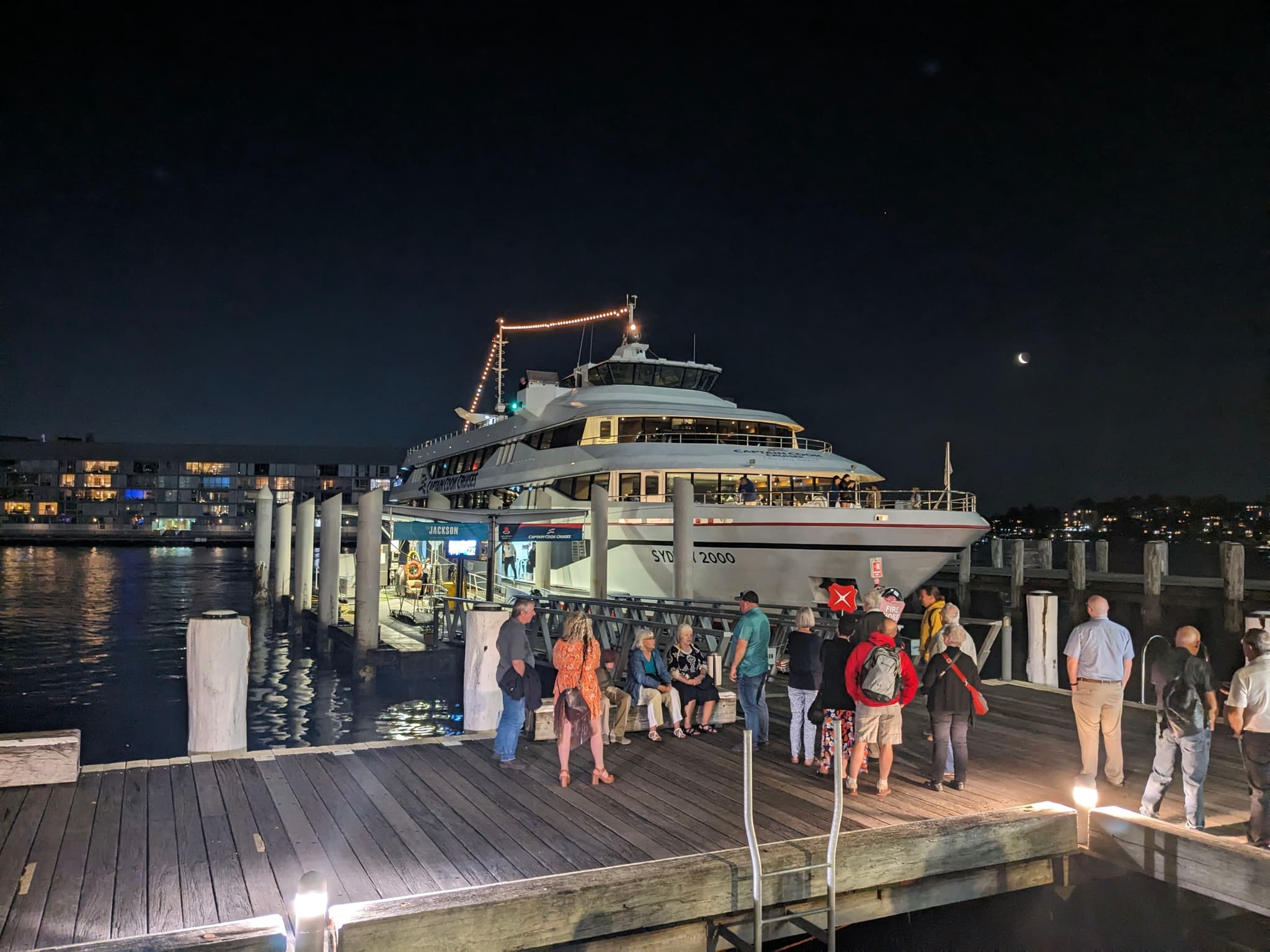 A group of people stands on a dock at night, with a large, illuminated boat moored nearby