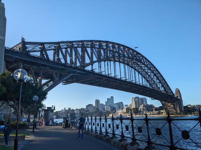 A large steel arch bridge spans across a body of water, with a city skyline in the background and a walkway with people and lampposts in the foreground