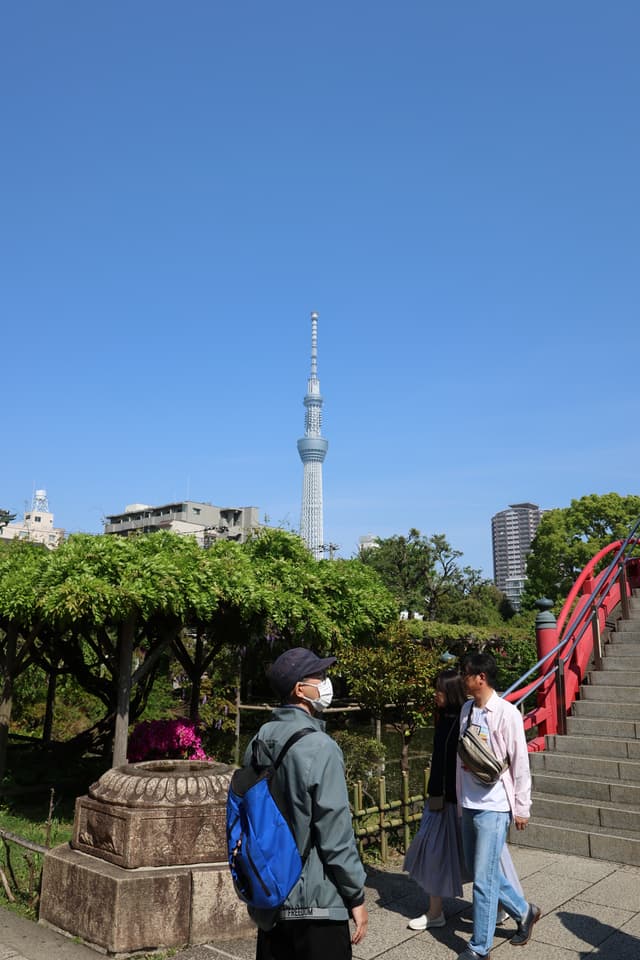 A tall tower rises in the background under a clear blue sky, with people walking near a red arched bridge and lush greenery in the foreground
