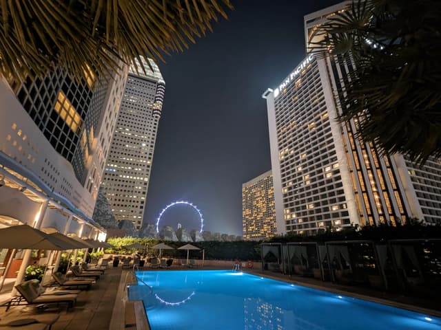 A nighttime cityscape featuring a brightly lit swimming pool surrounded by tall buildings, with a large Ferris wheel visible in the background