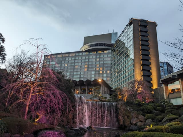 A tall, modern building with large windows is set against a cloudy sky. In the foreground, a landscaped garden features a waterfall and trees illuminated with pink lights