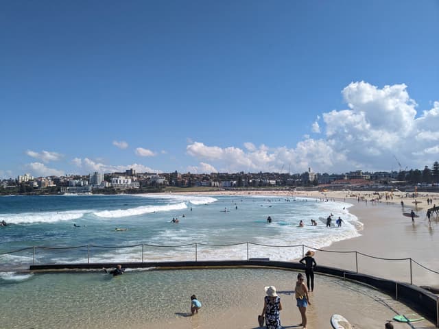 A sunny beach scene with people enjoying the water and sand, featuring a clear blue sky and scattered clouds