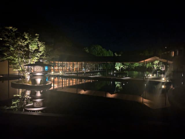 A serene nighttime scene featuring a well-lit building with large windows, surrounded by water and trees, reflecting lights on the water's surface
