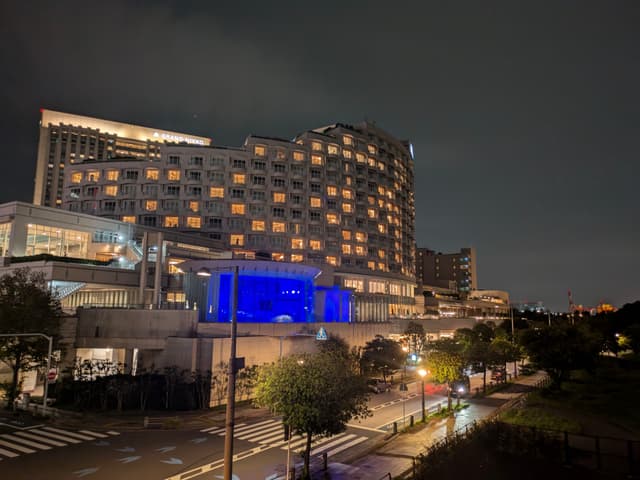 A large, illuminated building at night with many windows, featuring a prominent blue-lit section. The surrounding area includes trees and a road with streetlights