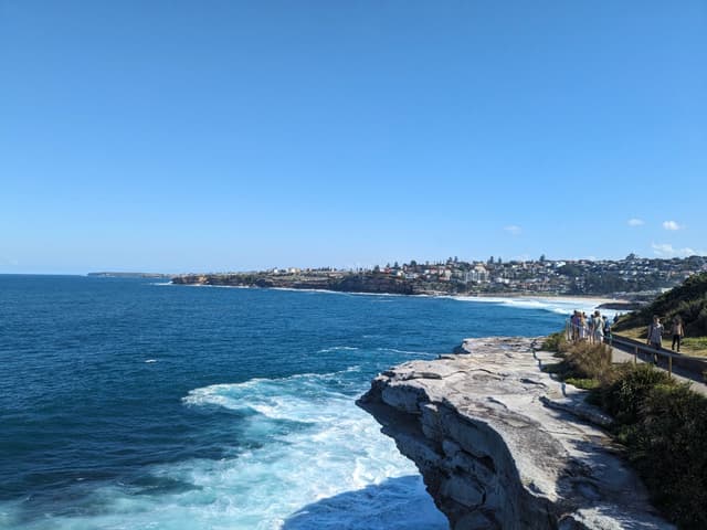 A coastal landscape with a rocky cliff overlooking the ocean, a clear blue sky, and a distant view of a shoreline with buildings