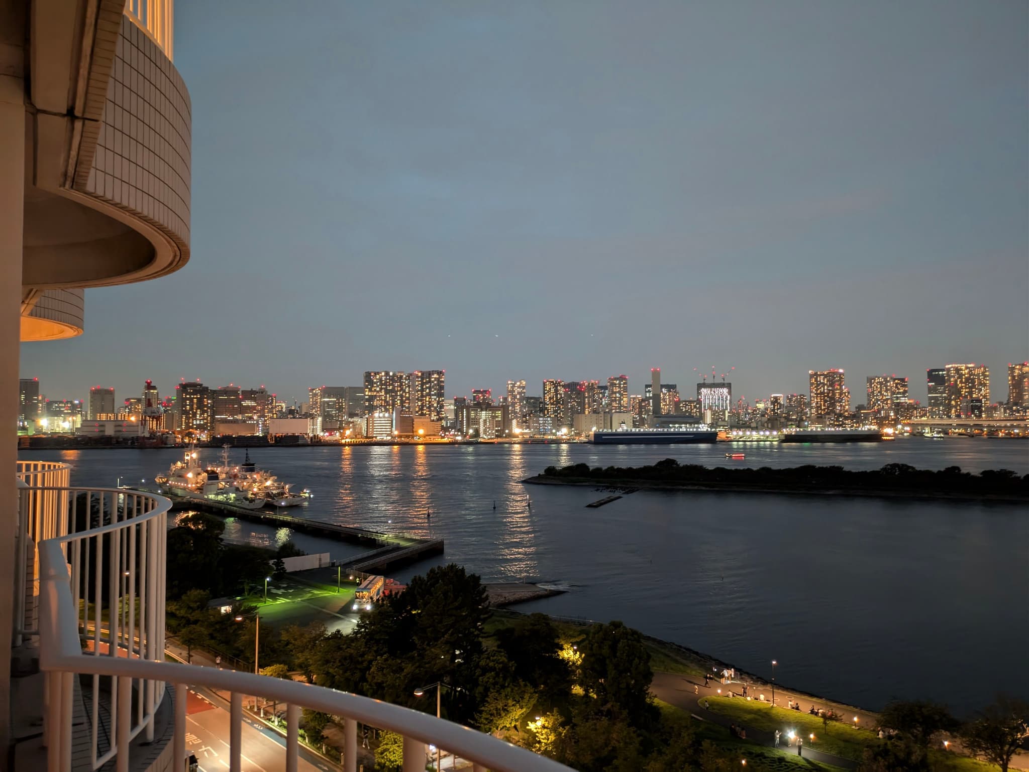 A cityscape at dusk with illuminated buildings across a body of water, viewed from a balcony